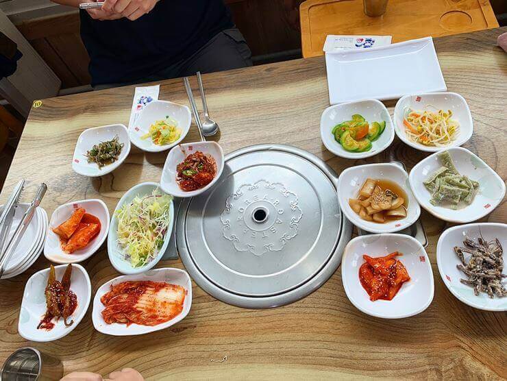 식탁 가운데 동그란 은색 쟁반을 중심으로 양 옆에 다양한 반찬이 놓여있다.