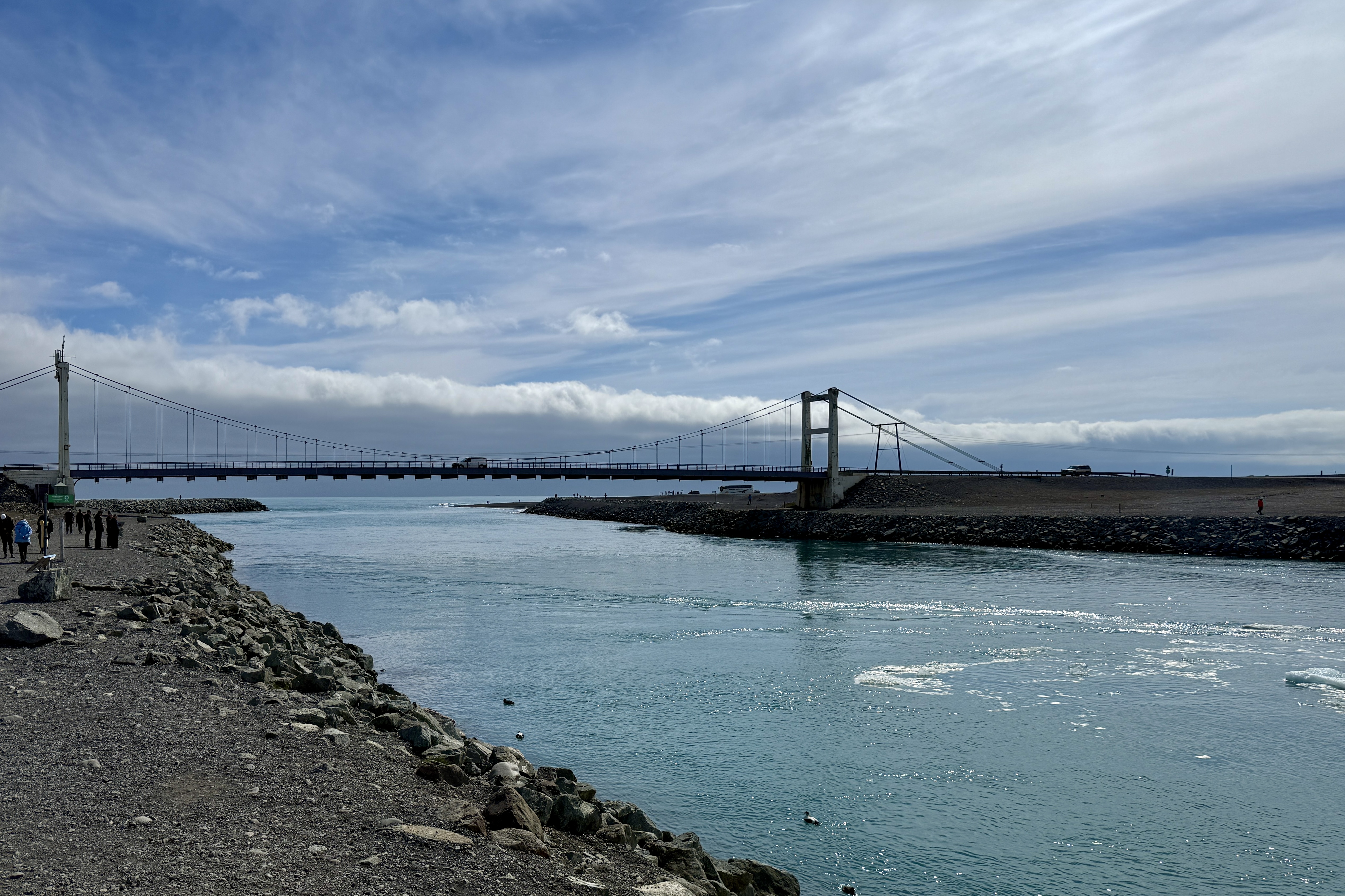 요쿨살론 호수와 다이아몬드 해변을 연결하는 다리. 청록색 물이 흐르는 수로 위에 가로질러 놓인 다리가 멀리 있고, 주변은 검은 자갈 해변으로 둘러싸여 있음. 맑은 하늘과 구름이 인상적이며, 물 위로 햇빛이 반사되어 반짝임.