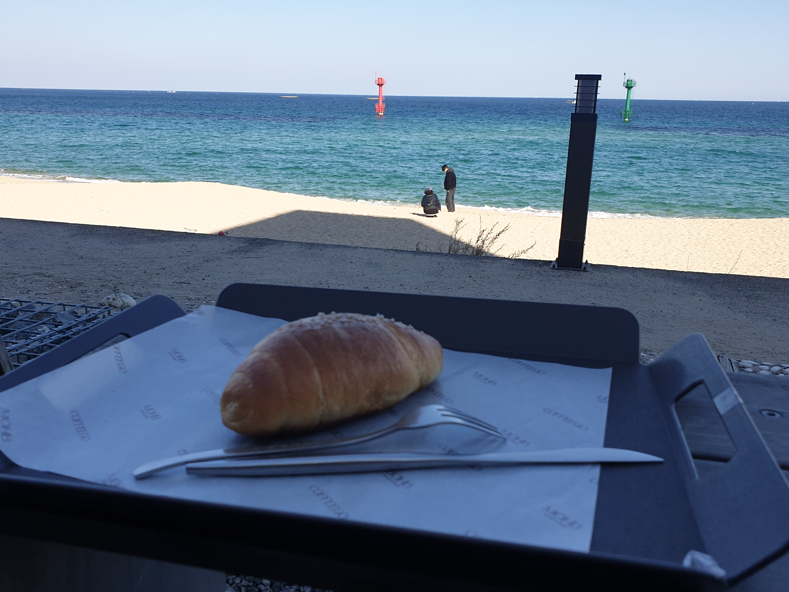 바다와 해변. 해변엔 두 사람이 있다. 해변을 바라보고 빵이 트레이에 올려져있다.