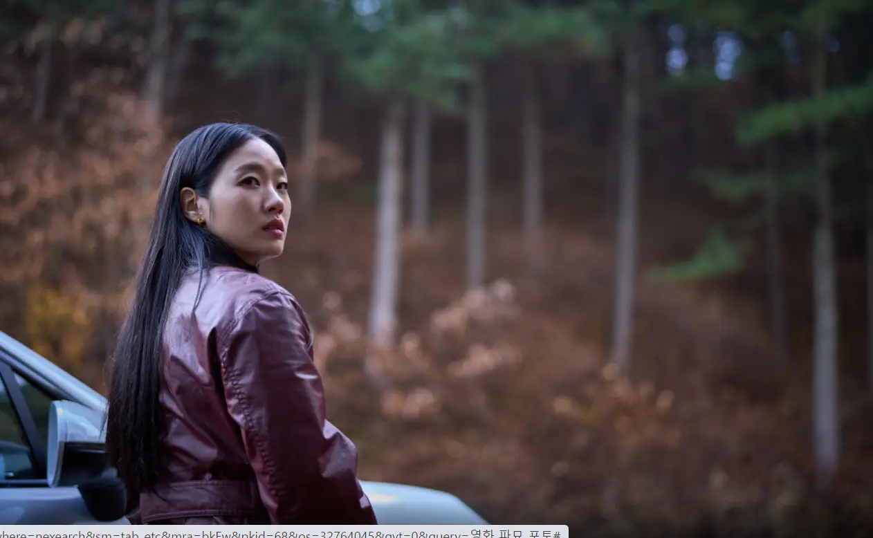 파묘- 숲이 빼곡한 산속 흰차 앞에서 빨간색 가죽옷에 긴머리를 늘어뜨리고 의심의 눈으로 옆을 바라보근 김고은