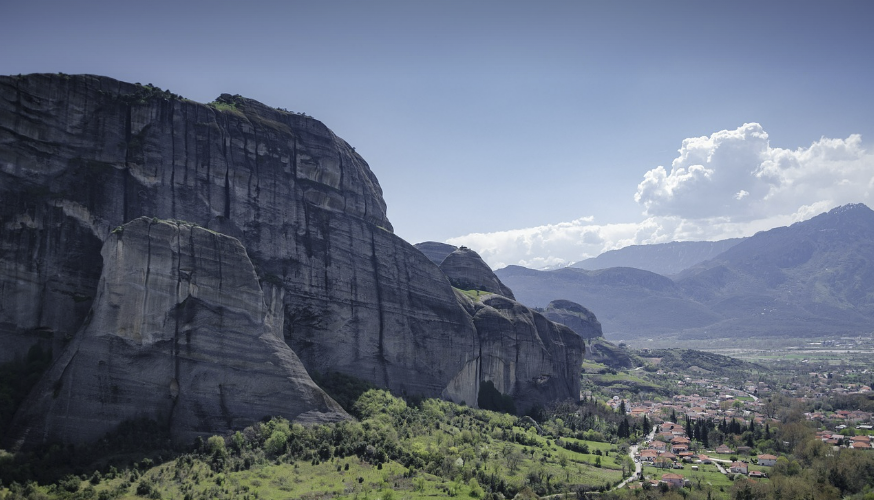&quot;그리스 메테오라의 거대한 바위 절벽과 그 아래 펼쳐진 작은 마을 풍경. 푸른 하늘 아래 드라마틱하게 솟은 암석과 멀리 보이는 산맥이 어우러진 전경&quot;