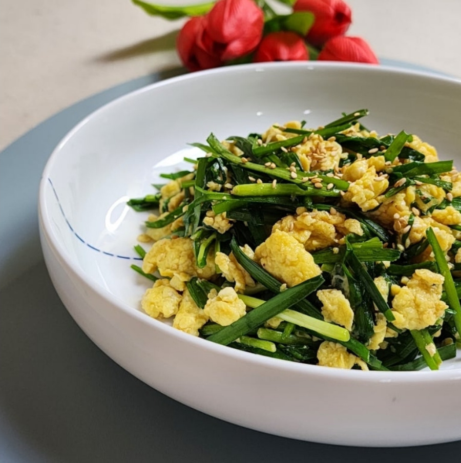 야생에서 채취를 한 부추를 볶은 후에 계란을 넣어 같이 버무린 후에 참깨를 뿌려서 동그랗고 하얀 그릇에 가득 담아두고 그 위에 장미꽃 5송이를 놓아두고 찍은 사진