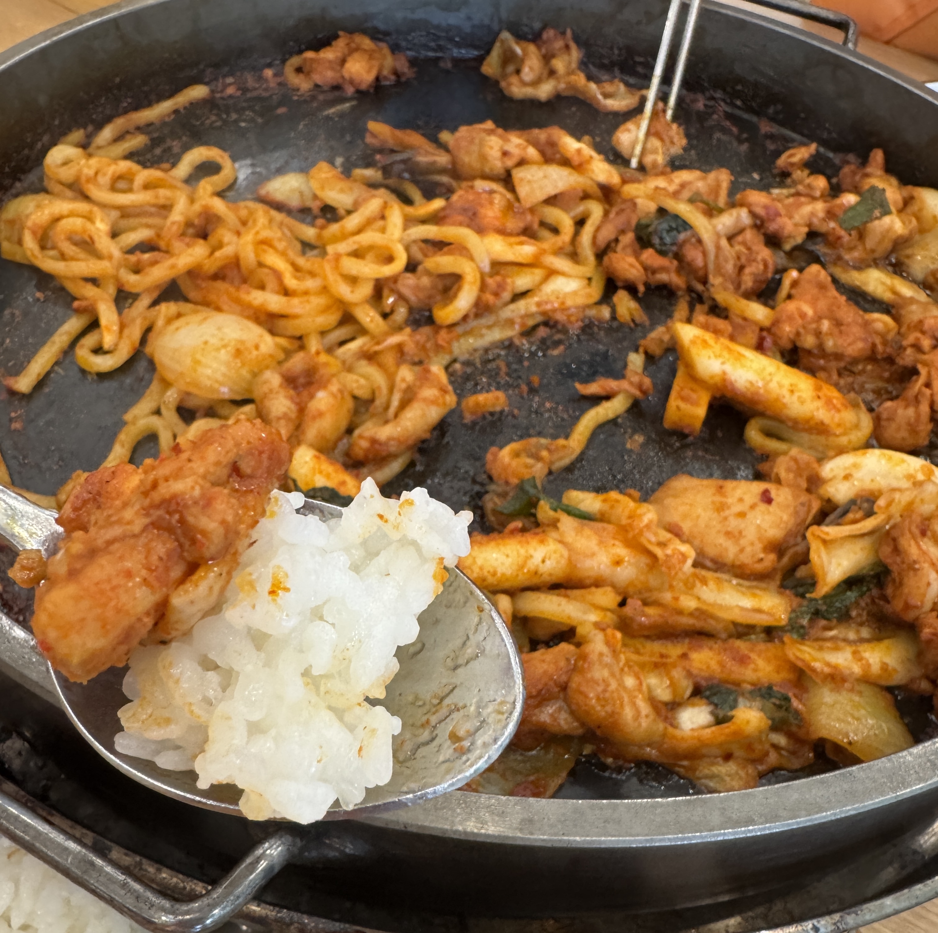 하얀 공깃밥 위에 매콤하게 양념된 닭갈비 한 조각을 올려 숟가락으로 든 모습. 뒤에는 우동사리와 닭갈비가 남아 있는 철판이 보인다.