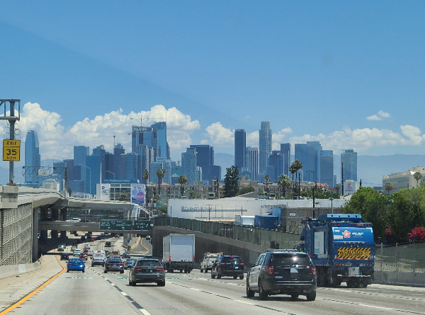 사진 아래쪽으로는 고가 차도 아래로 내려가는 차들. 위쪽으로는 다운타운의 높은 빌딩들이 한 곳에 모여 있는 모습들. 빌딩 뒤편에 하얗고 큰 뭉게구름이 있다.