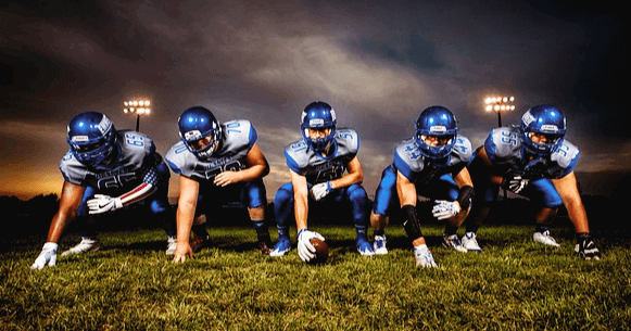 잔디밭에 6명의 남자 럭비선수들이 앉아있다. 파란 유니폼과 파란 헬맷을 썼다.