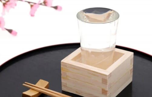나무 상자안에 유리로 된 잔이 들어가 있고 유리잔안에는 사케가 가득 따라져 있다.