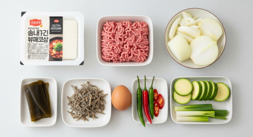 순두부찌개에 사용될 순두부, 다진 돼지고기, 달걀, 양파, 호박, 대파, 멸치와 다시마 등 모든 재료가 깔끔하게 정리된 모습의 평면 사진.