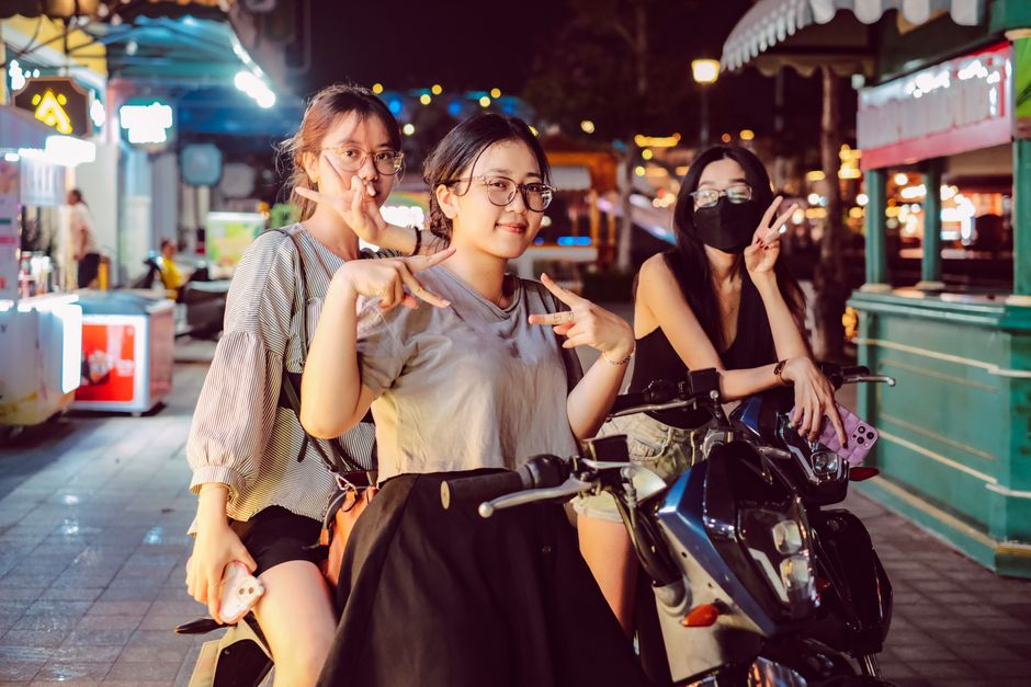 Three friends pose on scooters at a lively night market, showcasing urban nightlife.