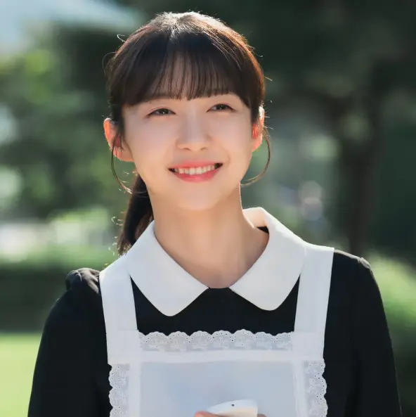 밝은 한낮 야외 정원에서 검은색 원피스에 흰색 앞치마를 두르고 정면을 바라보며 웃고있는 사월