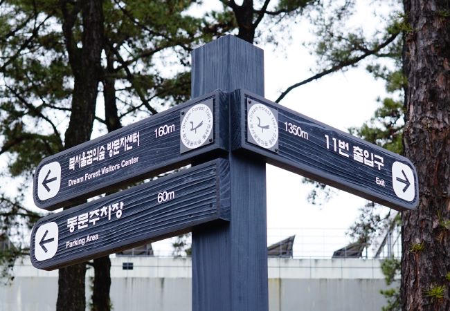 길안내 이정표&#44; 진한 회색ㅂ탕에 흰글씨&#44; 뒤로는 큰 나무&#44; 주차장 건물&#44; 나무 사이로 보이는 흰 하늘&#44;