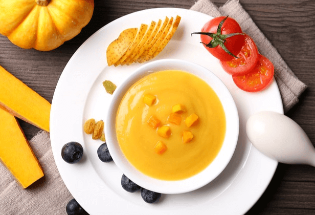하얀 그릇에 노란 호박죽이 가득 담겨있다. 왼쪽에는 둥그런 노란 호박이 있고 죽그릇 주변에는 블루베리들과 잘린 방울토마토 조각들이 있다.