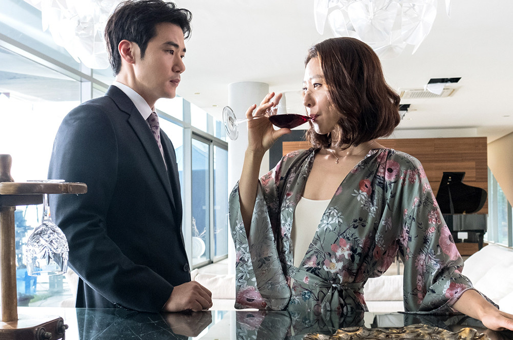 통유리가 있는 고급 저택 실내에서 정장 차림의 남자가 서 있고, 맞은편에 플로럴 가운을 입은 여자가 레드 와인을 마시며 그를 바라보고 있다