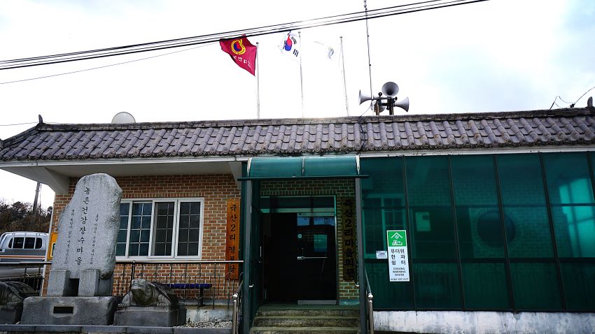 붉은 벽돌 건물, 죽산2리 마을회관, 회색 기와, 지붕에 좌 우 양방향의 스피커, 하얀 하늘에 태극기와 국기봉,