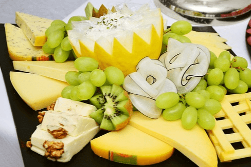매우 넓은 검정 쟁반에 다양한 치즈 조각들이 가득하다. 하얀 치즈, 노란 치즈 슬라이스들과 청포도들이 그 위에 놓여있다.