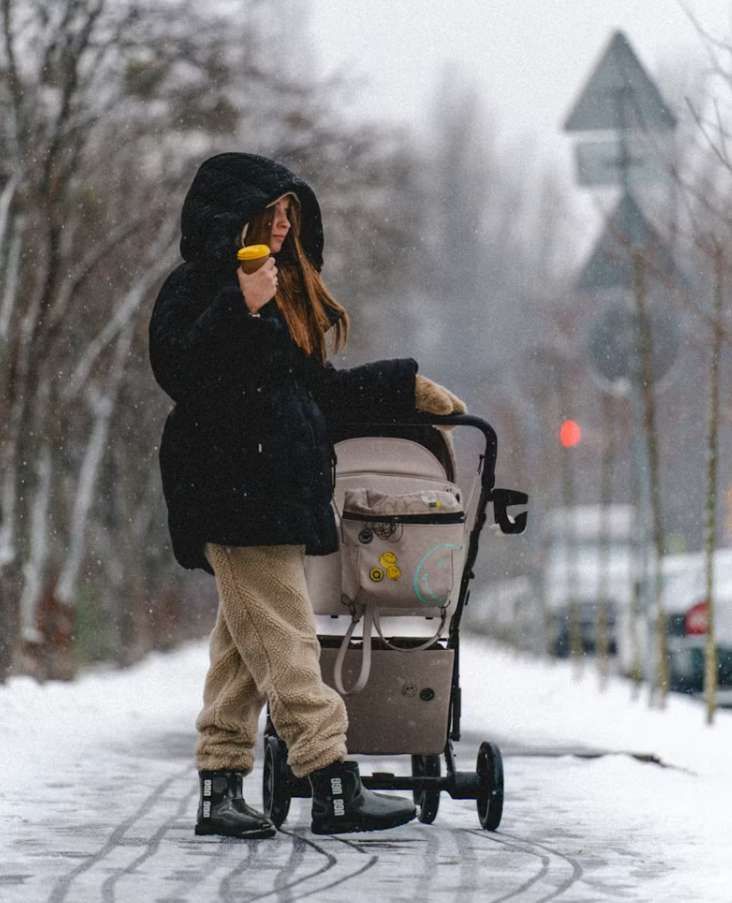 눈 내리는 겨울 거리에서 유모차를 밀며 따뜻한 음료를 들고 있는 여성