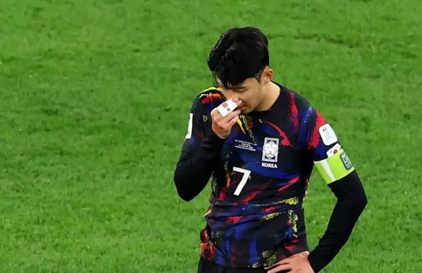 손흥민-
잔디밭 위 검은색과 붉은색 파란색이 섞인 유니폼을 입고 붕대를 감은 오른손 손가락으로 코를 잡으며 땅을 바라보는 손흥민