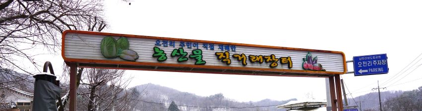 오전리 직거래장터 입구, 높게 걸어놓은 안내문, 회색 바탕에 녹색, 주황색 글씨, 하얀 하늘,