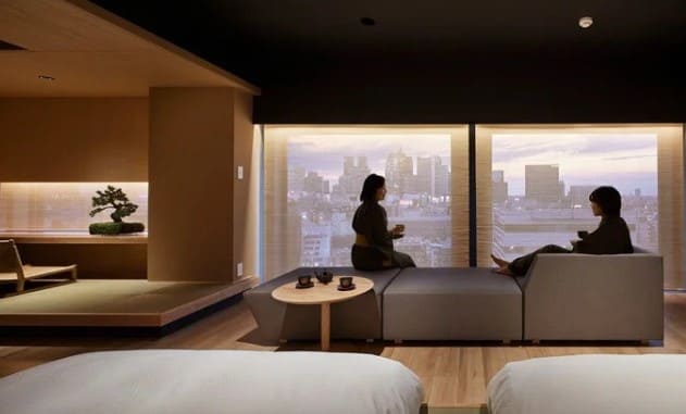 창 밖으로 빌딩들이 보이고 남자와 여자가 소파에 앉아서 서로를 바라보고 있다.