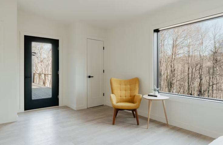 햇살이 드는 깔끔한 미니멀 인테리어 공간. 노란색 포인트 의자와 작은 사이드 테이블이 큰 창 옆에 놓여 있고, 창밖으로는 겨울 나무들이 보인다.