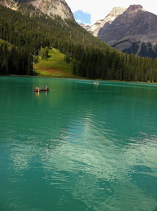 카누를 타고 에메랄드빛 호수를 가로지르는 사람들, 숲과 산자락이 병풍처럼 둘러싸인 풍경.