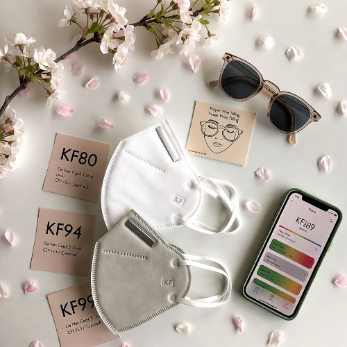 An editorial flat lay photograph on a clean white surface showing three KF masks neatly arranged &mdash; KF80, KF94, and KF99 &mdash; each with a small label card beneath. Beside the masks: a pair of simple clear-framed sunglasses, a small illustration card showing proper nose wire fitting technique, and an open smartphone showing an air quality app. Cherry blossom petals are scattered naturally around the scene. The mood is organized, practical, and spring-appropriate. Soft natural light from above. Warm neutral tones with soft sage green accents.