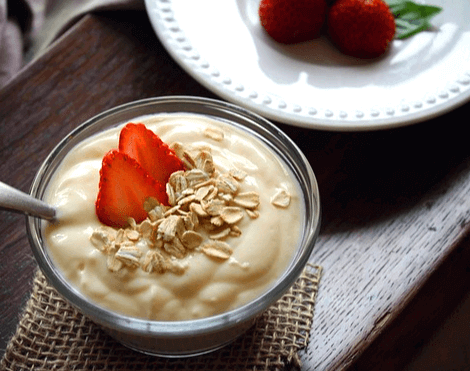 투명한 유리 그릇에 하얀색의 요구르트가 가득하고 그 위에는 빨간 딸기 조각 2개와 시리얼이 있다.