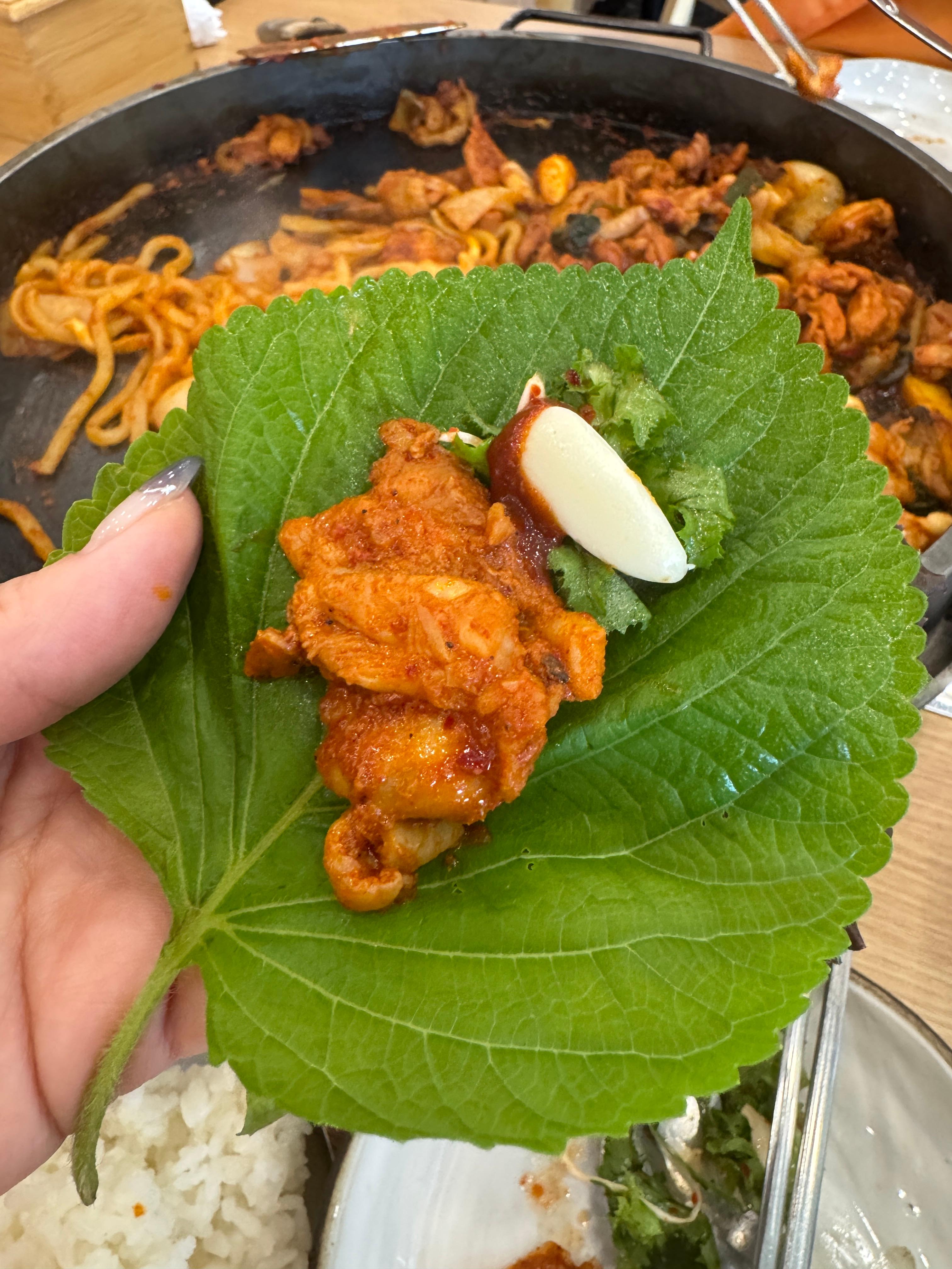 깻잎 위에 닭갈비와 마늘&amp;#44; 고추장 양념을 올려놓은 모습. 뒷배경에는 닭갈비 철판이 보인다. 