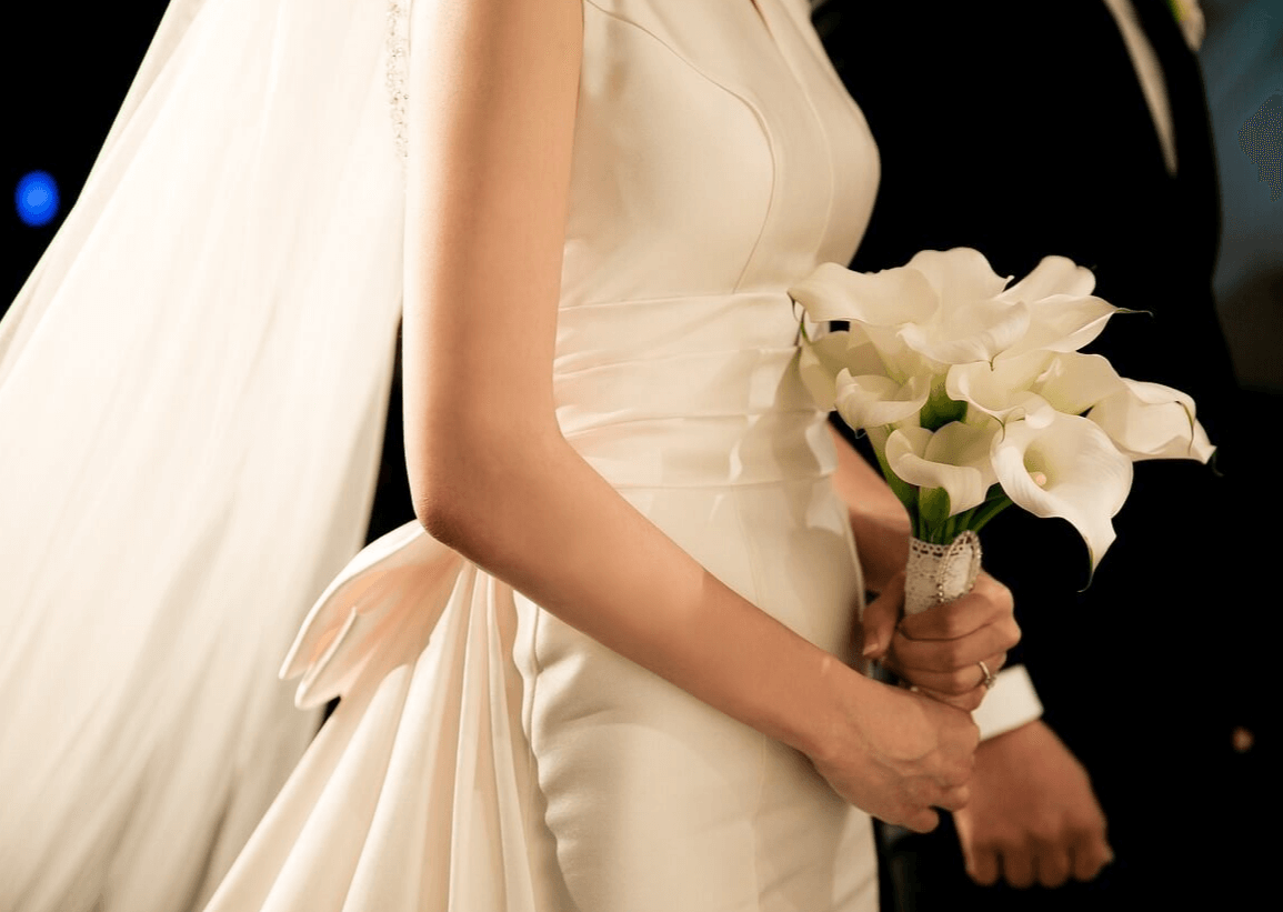 하얀 웨딩드레스를 입은 신부가 하얀 카라꽃을 들고 있고검정 양복을 입은 신랑과 함께 서 있다.