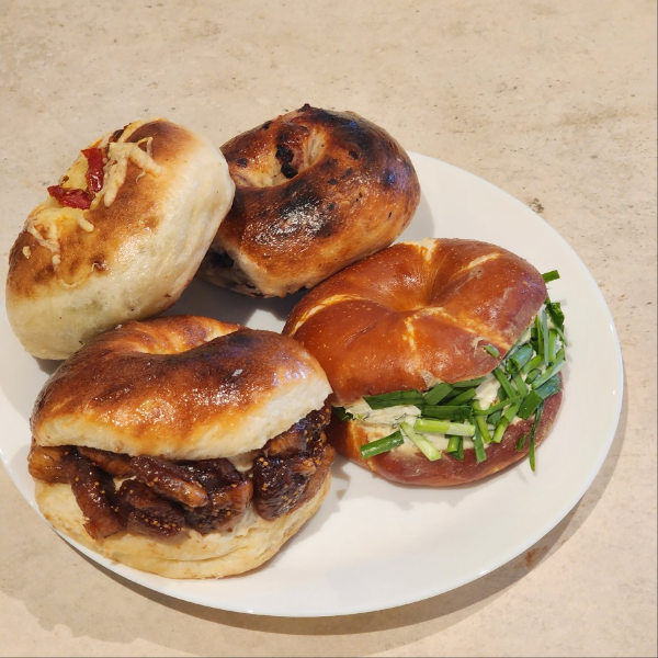 흰색 접시에 다양한 종류의 베이글과 베이글 샌드위치가 놓여 있음. 무화과와 크림치즈가 들어간 베이글, 부추와 크림치즈가 들어간 브레첼 베이글, 그리고 구운 베이글과 치즈가 토핑된 베이글이 함께 놓인 모습