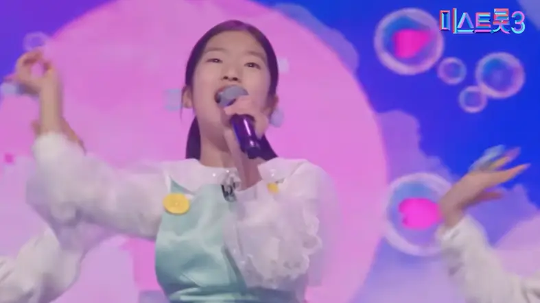 미스트롯3- 파란 배경 앞 하늘색 드레스를 입고 왼손으로 마이크를 들고 오른손으로 하늘을 가리키며 노래하는 빈예서