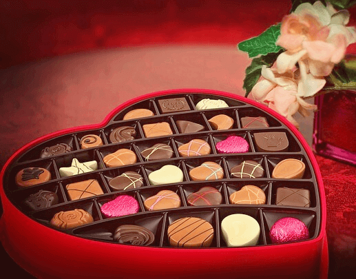 하트 모양의 빨간 상자 안에 20개의 예쁜 초콜릿이 담겨있고 오른쪽에는 분홍장미가 있는 꽃병이 있다.