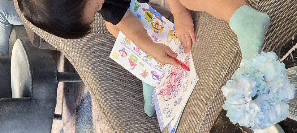 소파에 앉아 뽀로로 그림책 위에 빨간색 색연필로 그림을 그리며 몰입해 있는 아기의 모습