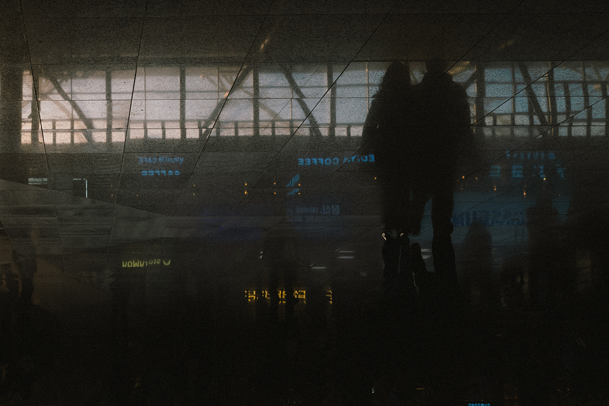 도로 위에 햇빛이 반사되어 그림자가 만들어져있다. 그 그림자 위로 사람들이 걸어가는 모습과 가게의 간판이 반사되어있다.