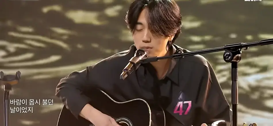 싱어게인3- 회색물결배경 검은 셔츠 가슴에 47번호표를 달고 검은색 기타를 치며 스탠딩마이크를 앞에 두고 두눈 감은채 노래하는 47호 