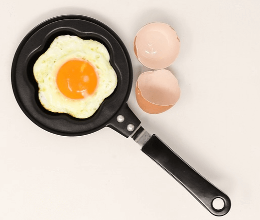 아주 작은 검정색 프라이팬에 노른자가 또렷하게 있는 달걀 프라이가 있다. 오른쪽에는 반으로 나뉜 달걀 껍질이 있다.