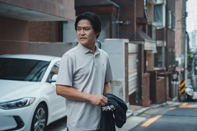 어느 주택 골목을 배경으로 회색 반팔티를 입고 서있는 영화 타겟의 주형사