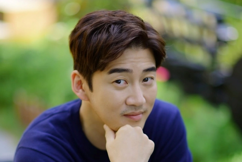 야외를 배경으로 갈색머리와 파랑 티를 입고 테이블에 앉아 있는 배우 윤계상