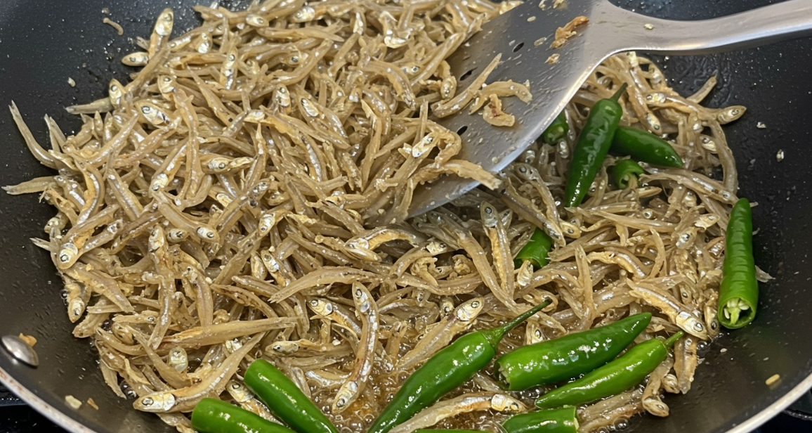 프라이팬에서 멸치가 노릇하게 볶이며, 청양고추가 살짝 투명해지는 모습. 주걱으로 부드럽게 섞고 있음.