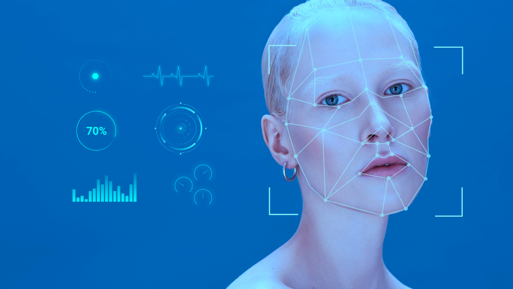 파란 배경에 흰색 피부를 가진 사람 얼굴이 나온다. 얼굴에 그물망 같은 선이 그려져 있고 얼굴 특징점을 연결해 매핑하는 모습이다. 왼쪽에는 심장박동 그래프, 70% 표시된 원형 그래프, 막대 그래프 등 다양한 데이터 시각화 요소가 배치되어 있다. 이는 얼굴 인식 기술이 작동하는 방식과 얼굴에서 데이터를 수집하는 과정을 보여준다.