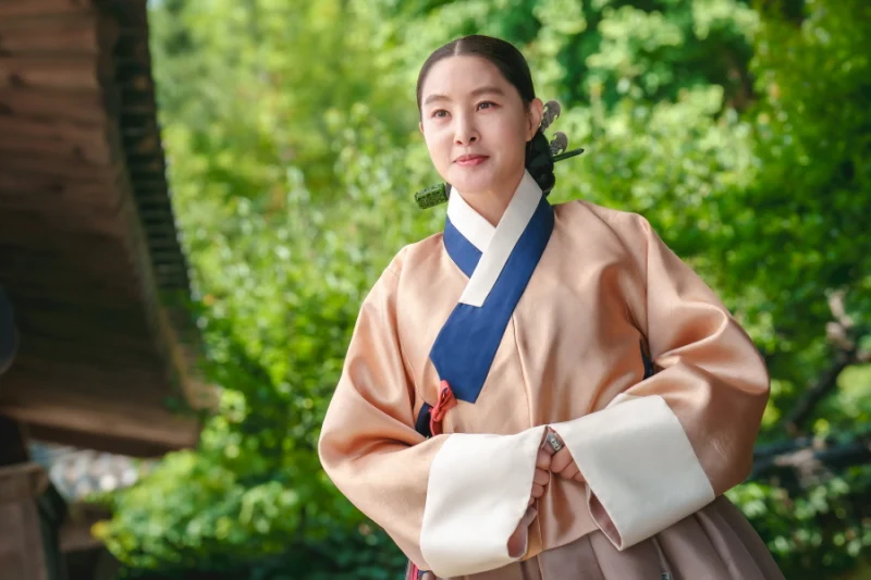 초목이 우거진 한옥을 배경으로 베이지색 한복을 입고 서 있는 사극 드라마 혼례대첩의 박소현 캐릭터