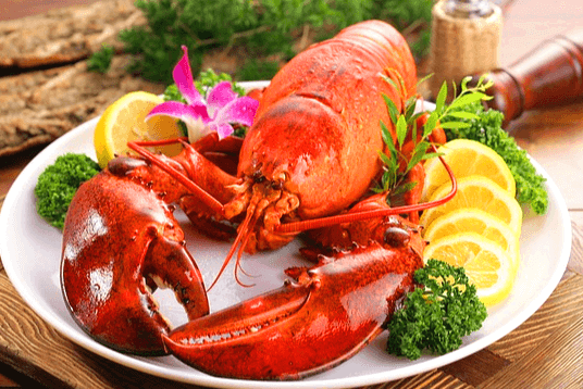 동그란 하얀 접시 위에 붉게 삶아진 커다란 랍스터 한 마리가 있고, 오른쪽에는 노란 레몬 조각 4개와 초록색 파슬리가 있다.