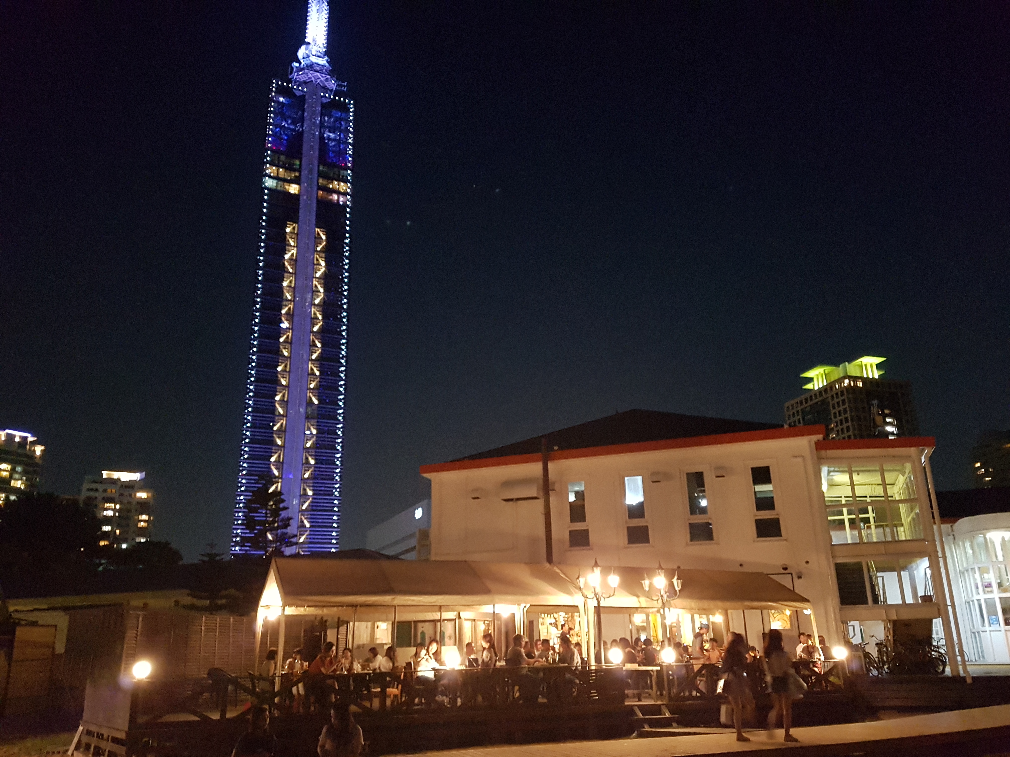 화려한 조명이 켜진 밤의 후쿠오카 타워와 그 아래 활기찬 해변 레스토랑 테라스의 야경.