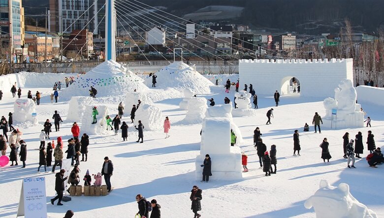 대관령 눈꽃축제장이 한눈에 내려다 보이는 전경사진으로 거대한 성벽에 출입구가 있고 사람들이 드나들고 곳곳에 커다란 눈조각품들이 전시되어 있는 곳으로 관광객들이 주변을 구경하고 사진을 찍고 있는 모습