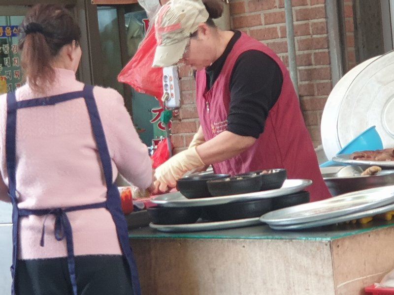 모자를 쓴 사장님이 순대를 썰어 뚝배기에 담고 있다. 직원 그 앞에서 대기하고 있다.