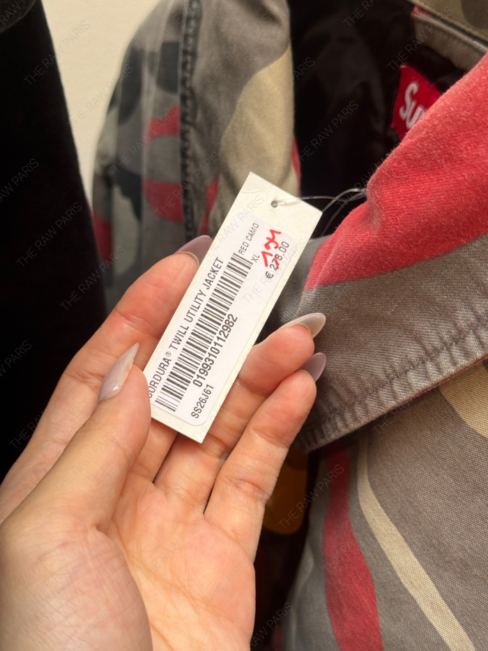 A close-up of a hand holding a Supreme Cordura Twill Utility Jacket price tag with hand-written discounted price of 139 euros⁠