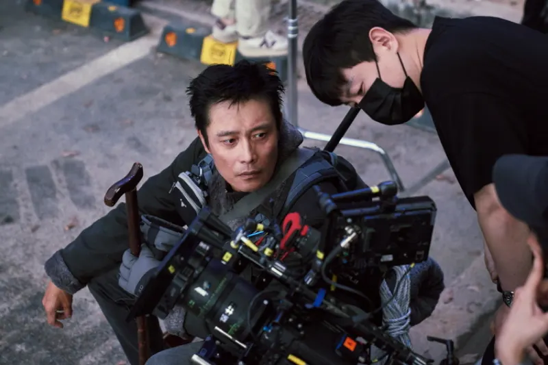 허름한 겨울 코트를 입고 지팡이를 쥔 채 촬영하고 있는 배우 이병헌