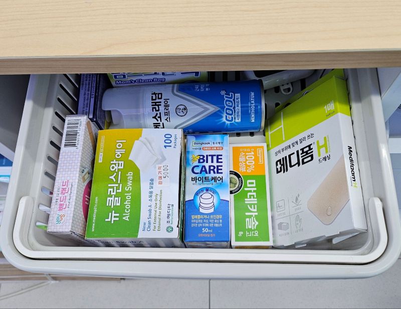 편의점 카운터 아래 서랍에 비치된 동네 아이들을 위한 비밀 응급 바구니. 뉴클린스왑, 마데카솔, 메디폼 H, 캐릭터 밴드 등이 정갈하게 담긴 모습.