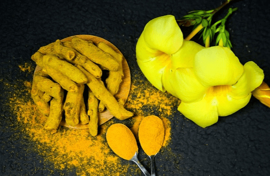 검정 식탁에 노란 강황이 가득하고 바닥에는 노란 강황가루가 흩어져있다. 오른쪽에는 노란 꽃이 있다.