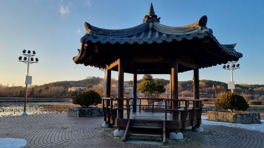 육각정자, 역광, 바닥에 둥근 원을 그리며 시공한 타일바닥, 왼쪽에 보안등, 가운데 정자로 올라가는 계단, 푸른 하늘,