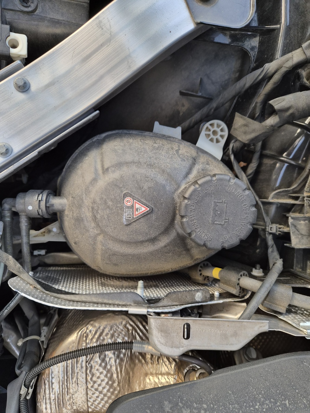 차량 보닛 안쪽에 위치한 냉각수 리저버 탱크 사진. 검은색 플라스틱 케이스와 ‘뜨거울 때 열지 말 것’ 경고 표시가 보임.